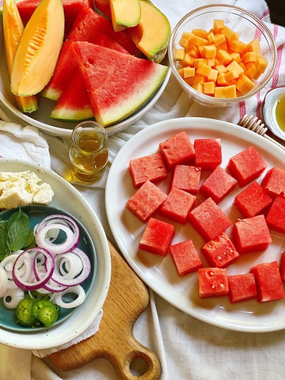 Ingredients for a watermelon salad, including watermelon cubes, sliced cantaloupe, cheese cubes, onions, mint leaves, and a small bottle of dressing.