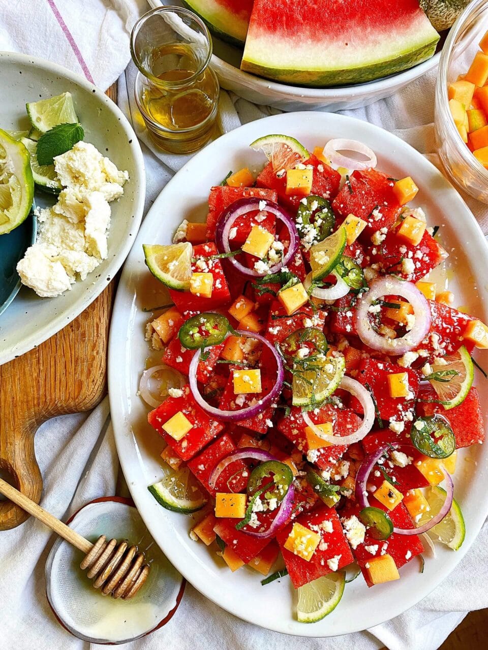 Plate of watermelon salad topped with cheese cubes, red onion rings, jalapeno slices, and lime wedges, surrounded by additional ingredients and dressing.