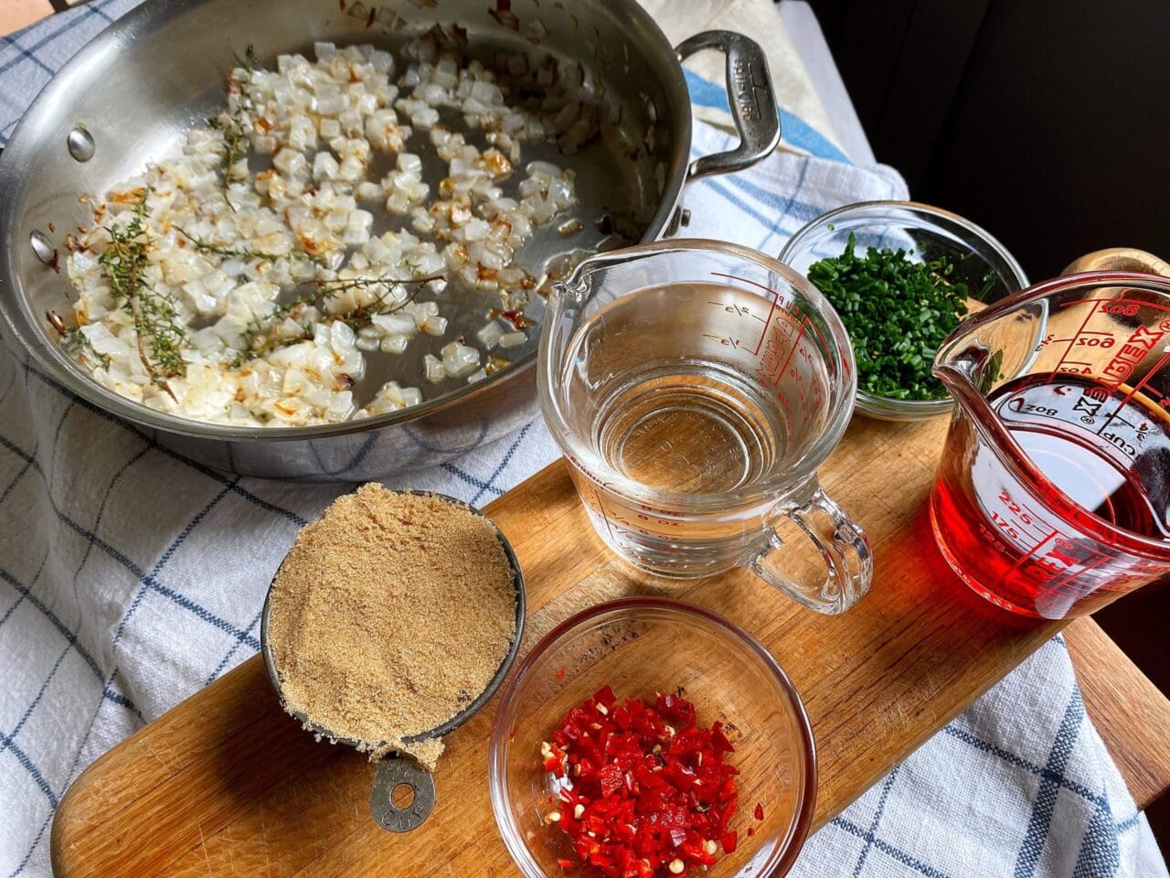 A cooking setup with a pan of sautéed onions and thyme, surrounded by ingredients on a wooden board: a cup of brown sugar, chopped red chilies, a bowl of chopped herbs, a measuring cup of water, and a measuring cup of red liquid.