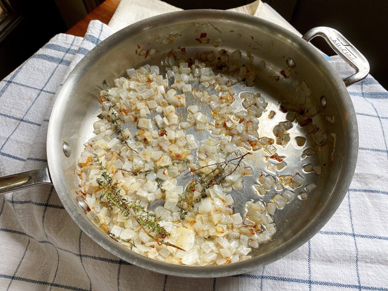 A stainless steel pan with diced onions and sprigs of thyme sautéing, placed on a checkered kitchen towel.