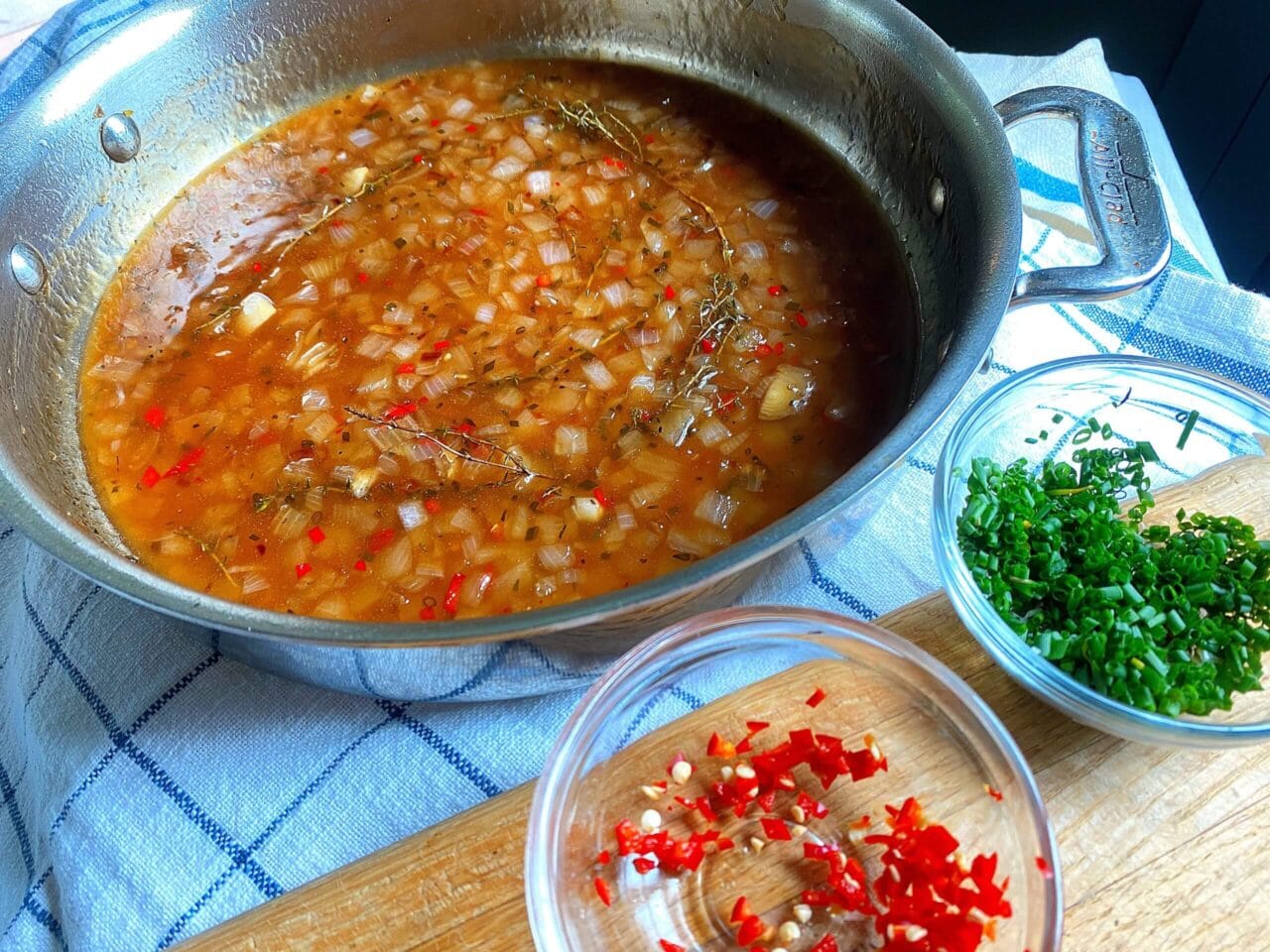 A pan of simmering sauce with onions, thyme, and red chilies, accompanied by bowls of chopped red chilies and green herbs on a wooden board.
