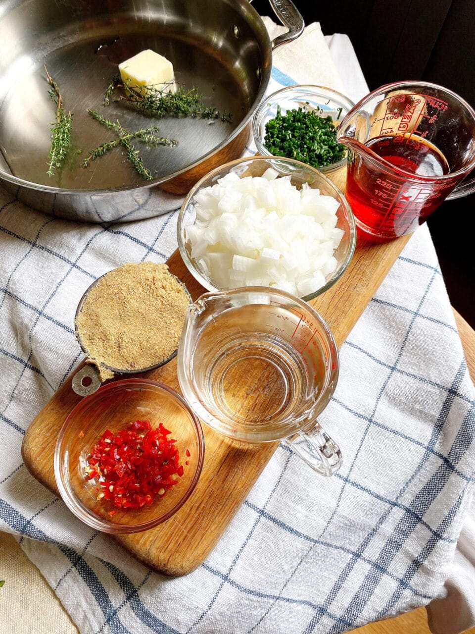 Ingredients for cooking including butter, thyme, chopped onions, chives, red pepper, brown sugar, water, and red liquid in a measuring cup on a wooden board.