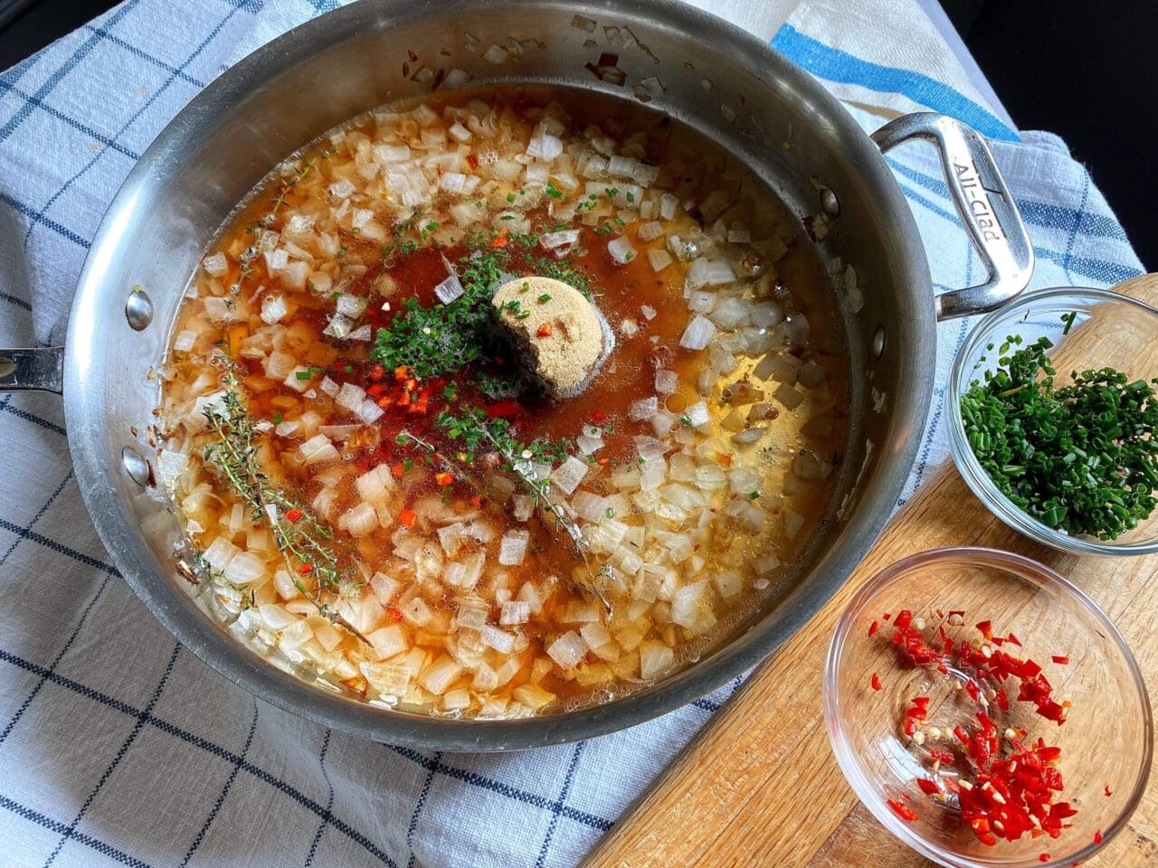 A pan filled with a cooking mixture of onions, broth, thyme, chopped red chilies, and a scoop of brown sugar, placed on a checkered kitchen towel with bowls of chopped herbs and chilies on a wooden board.