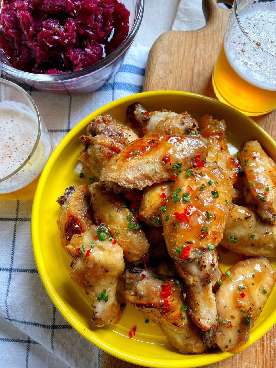A plate of glazed chicken wings garnished with chopped herbs and red chilies, accompanied by glasses of beer and a bowl of pickled red cabbage.