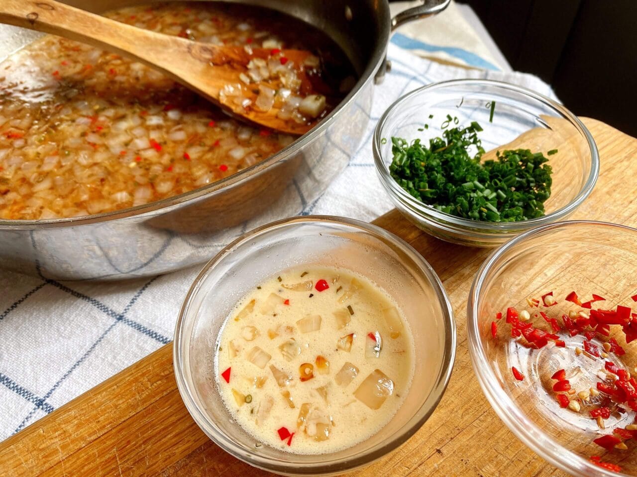 A pan of simmering broth with onions and red chilies, accompanied by bowls of chopped herbs, chopped red chilies, and a small bowl of creamy onion and chili mixture, all placed on a wooden board.