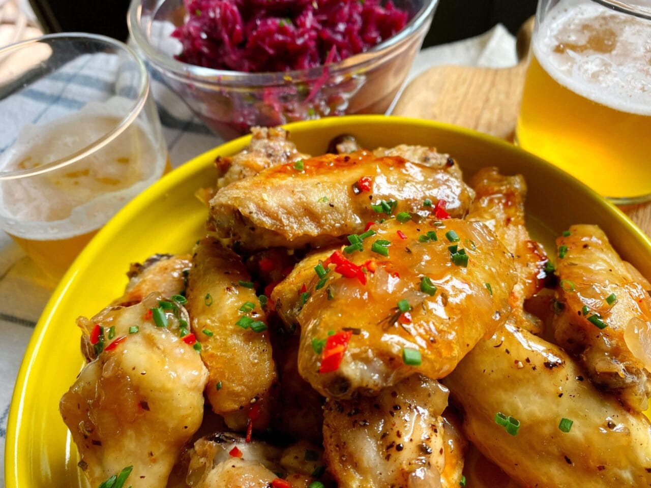 A close-up of glazed chicken wings garnished with chopped herbs and red chilies, accompanied by glasses of beer and a bowl of pickled red cabbage in the background.