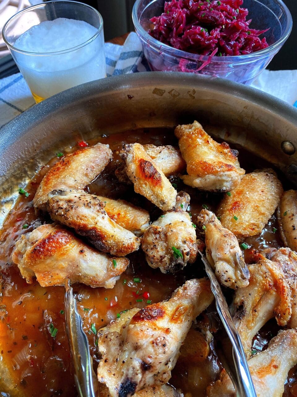 A pan of glazed chicken wings garnished with chopped herbs and red chilies, with a bowl of pickled red cabbage and a glass of beer in the background.