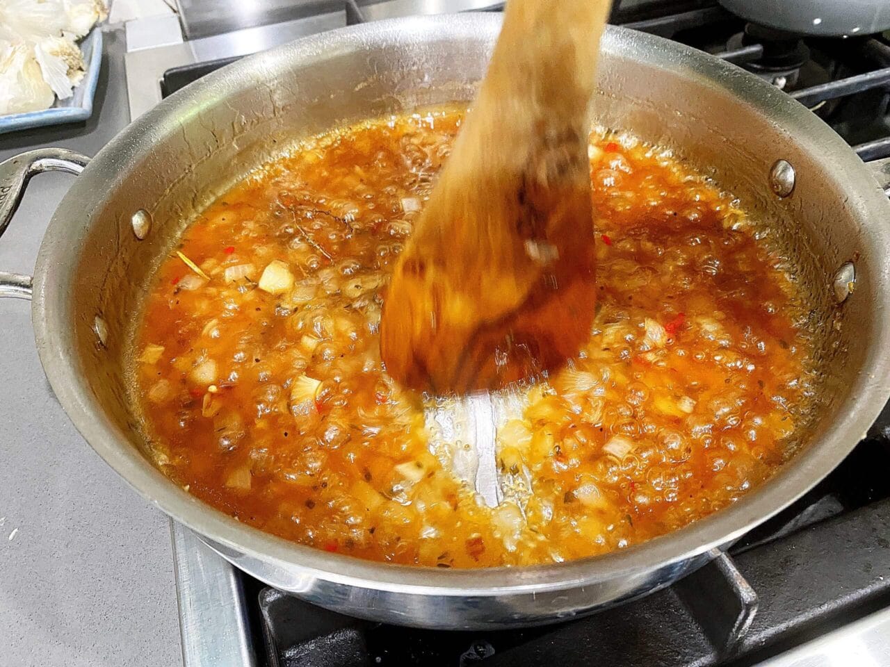 A pan of bubbling sauce with chopped onions and red chilies being stirred with a wooden spoon on a stovetop.