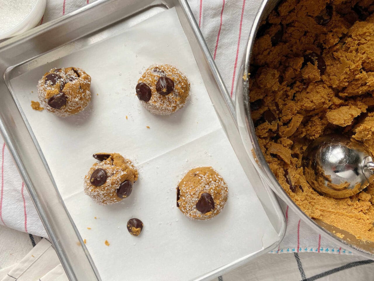 Overhead view of four cookie dough balls with chocolate chips and sugar on a parchment-lined baking sheet next to a bowl of cookie dough with a scoop.