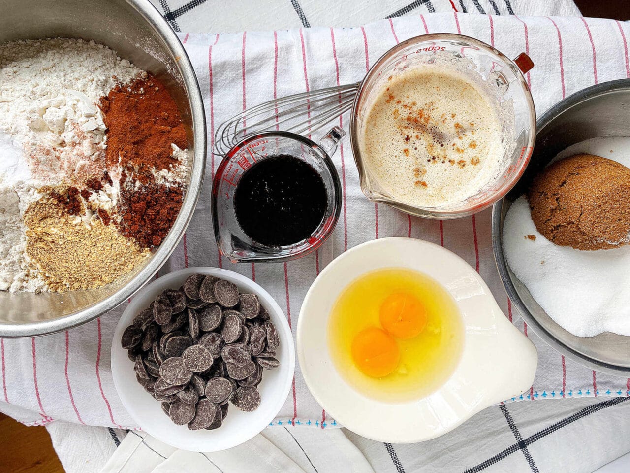 Overhead view of baking ingredients for cookies, including flour, spices, brown sugar, chocolate chips, eggs, and liquids in various bowls and measuring cups on a striped kitchen towel.