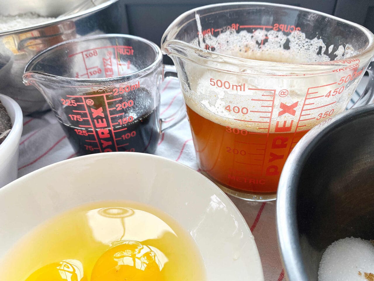 Close-up view of liquid ingredients for baking, including eggs in a bowl and measuring cups with melted butter and dark liquid, likely molasses, on a striped kitchen towel.