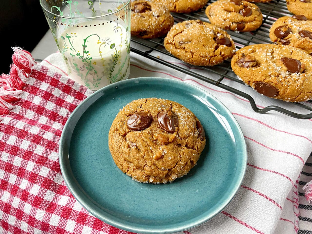 Chocolate chip cookie on a blue plate with a glass of milk and a red checkered napkin, with more cookies cooling on a wire rack in the background.
