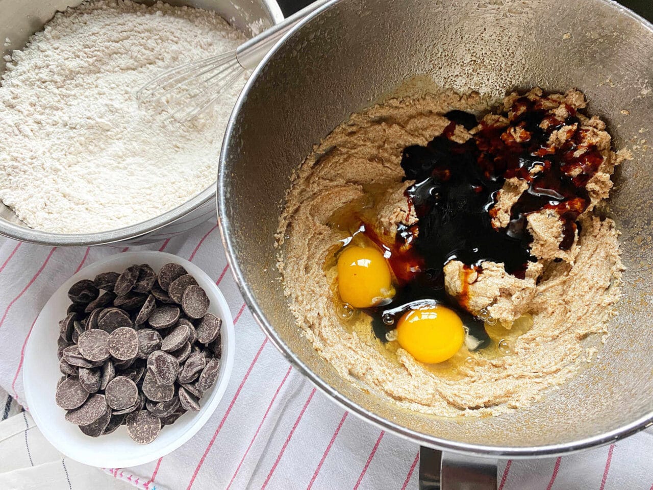 Mixing bowl with cookie dough ingredients, including eggs and molasses, next to bowls of flour and chocolate chips on a striped kitchen towel.