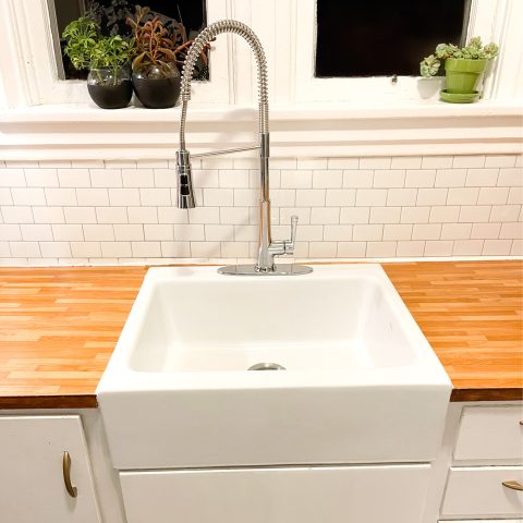 A glossy white rectangular Quick-Fit® drop-in farmhouse sink made in fireclay with a chrome faucet set in butcher block countertop and white cabinets with white tile backsplash.