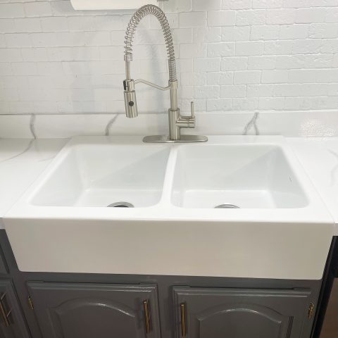 A glossy white rectangular Quick-Fit® double bowl drop-in farmhouse sink made in fireclay with a brushed stainless faucet set in gray cabinets in front of a white backsplash.