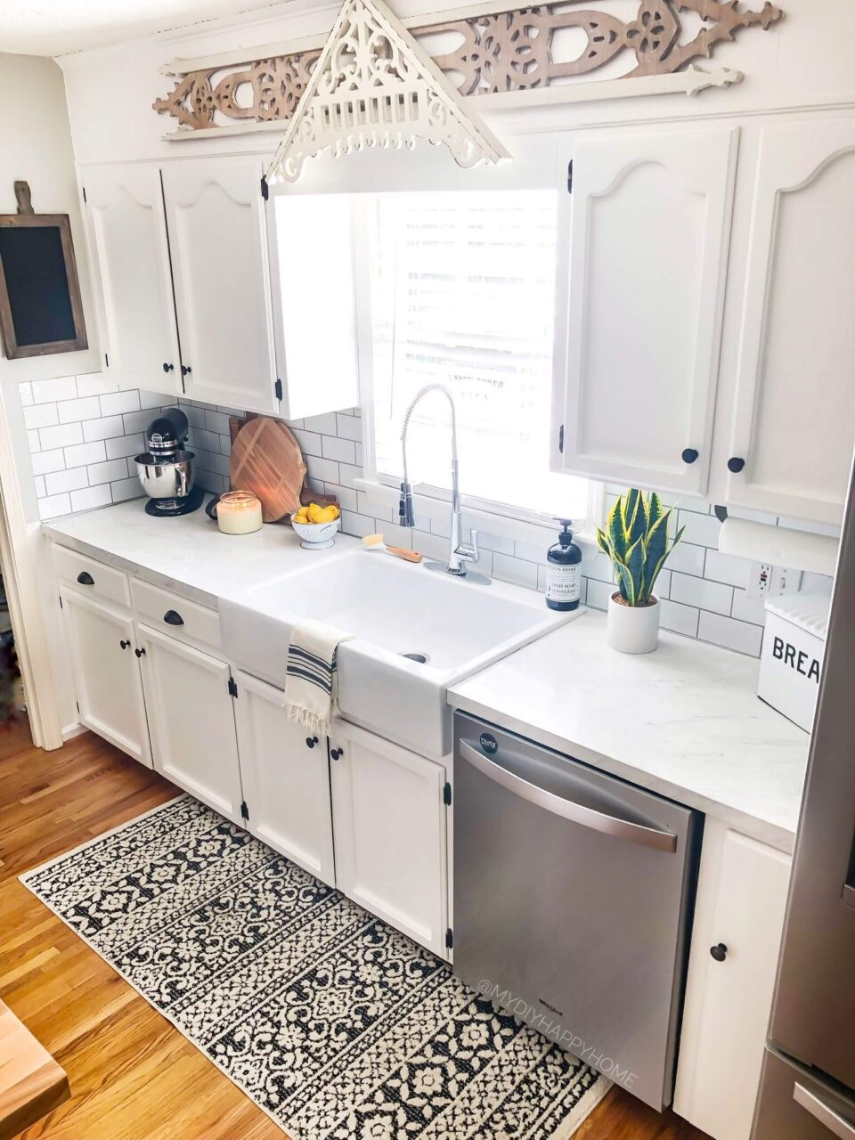 A bright, white kitchen with farmhouse-style decor, featuring a farmhouse sink, stainless steel dishwasher, and a patterned rug on a wooden floor.