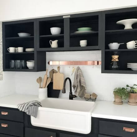Modern farmhouse kitchen sink with black open cabinets, white countertops, bronze faucet, and potted plants.