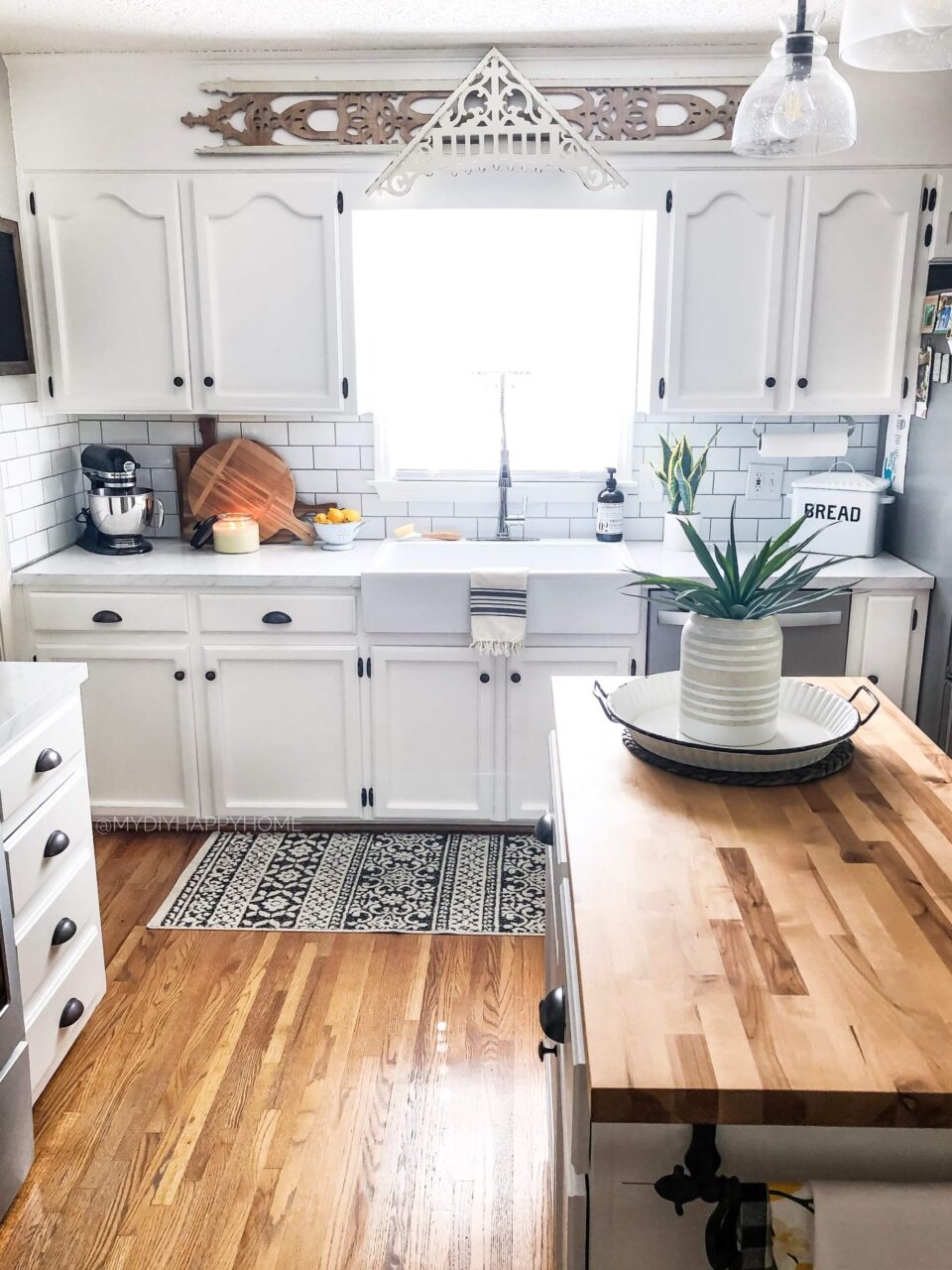 A farmhouse-style kitchen with white cabinets, a wooden island, and a decorative backsplash, featuring a patterned rug on a wooden floor.