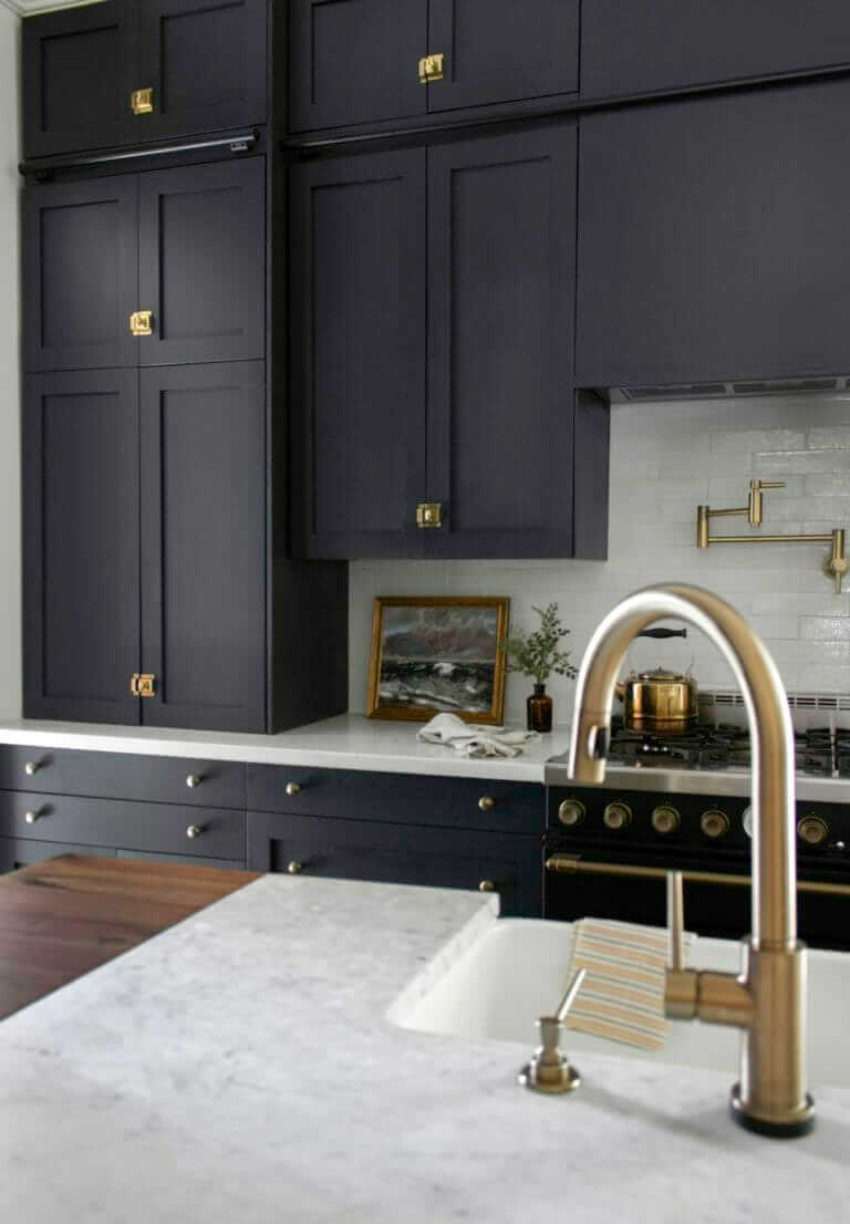 A modern kitchen with dark cabinetry, a white marble countertop, a brass faucet, and a framed painting placed on the counter near the stove.