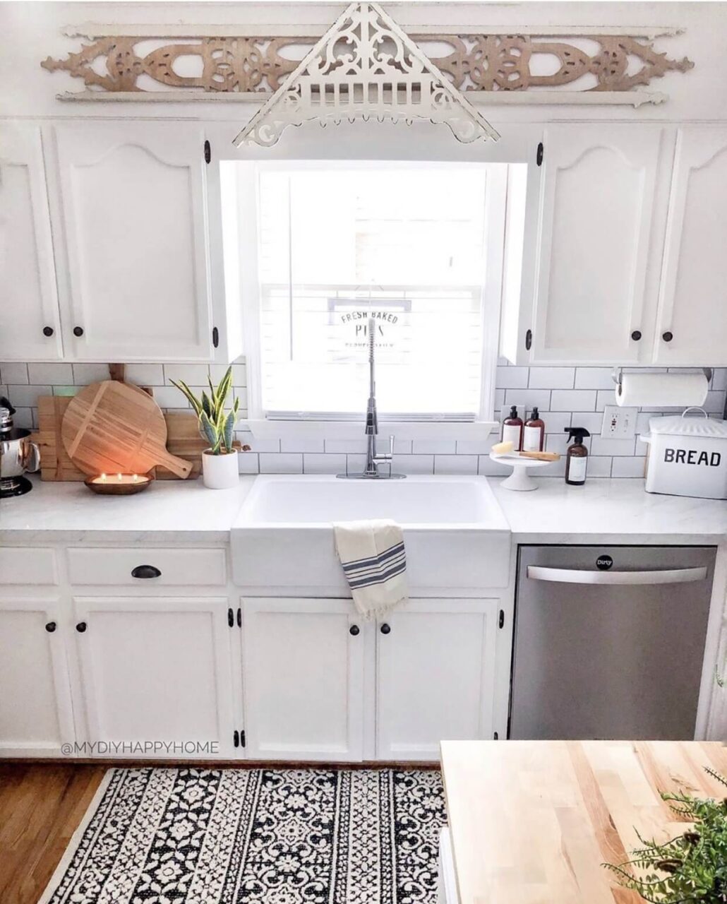 A cozy kitchen with white cabinetry, a farmhouse sink, and a patterned rug, featuring a decorative wooden trim above the window.