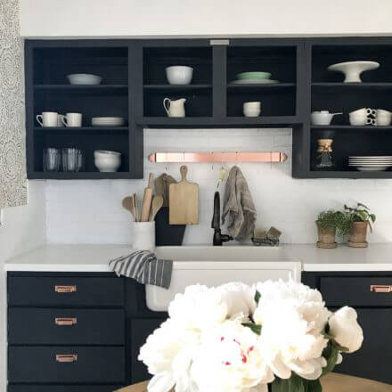 Modern kitchen with open black cabinets, white countertop, farmhouse sink, and a bouquet of flowers in the foreground.