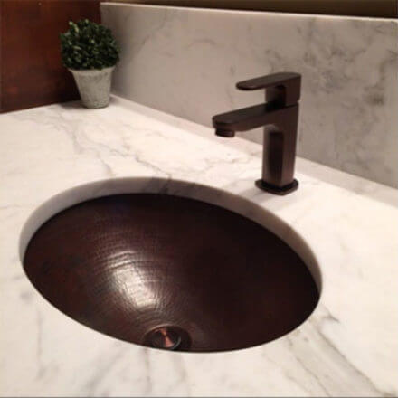 Modern bathroom vanity with a round copper sink set in a white marble countertop, paired with a sleek, dark faucet and a small potted plant in the background.