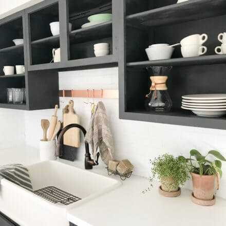 Modern farmhouse kitchen sink with black open cabinets, white countertops, bronze faucet, and potted plants.