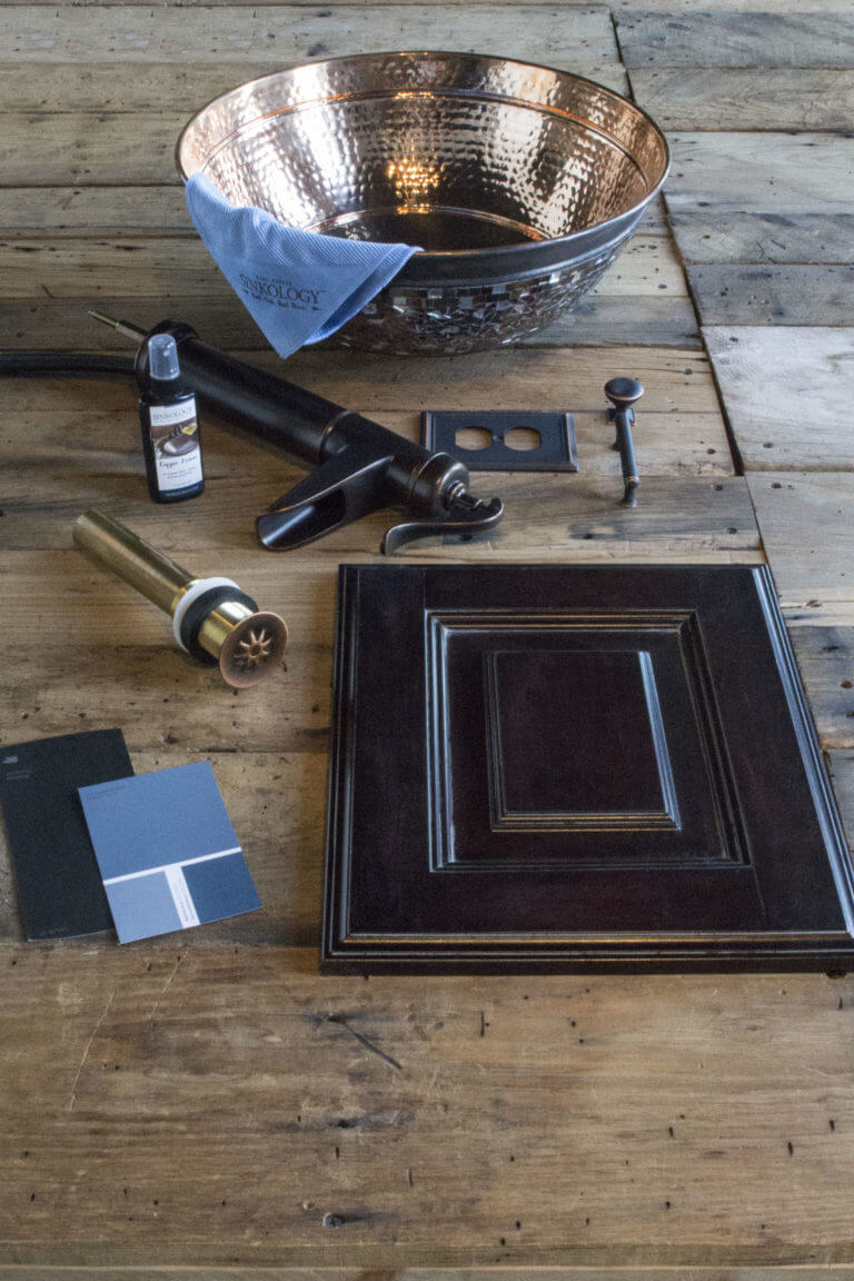 A hammered copper bowl sink with a mosaic exterior, a dark faucet, a cabinet door sample, and various hardware pieces on a wooden surface. The setup includes a blue cleaning cloth with the Sinkology logo and a bottle of "Copper Armor" cleaner.