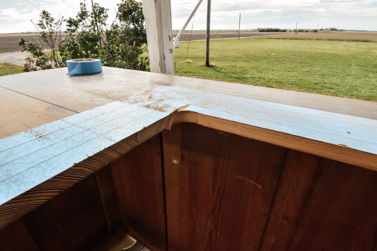A wooden surface with blue painter's tape along the edge, partially cut, with a roll of tape resting on top and a green field in the background.