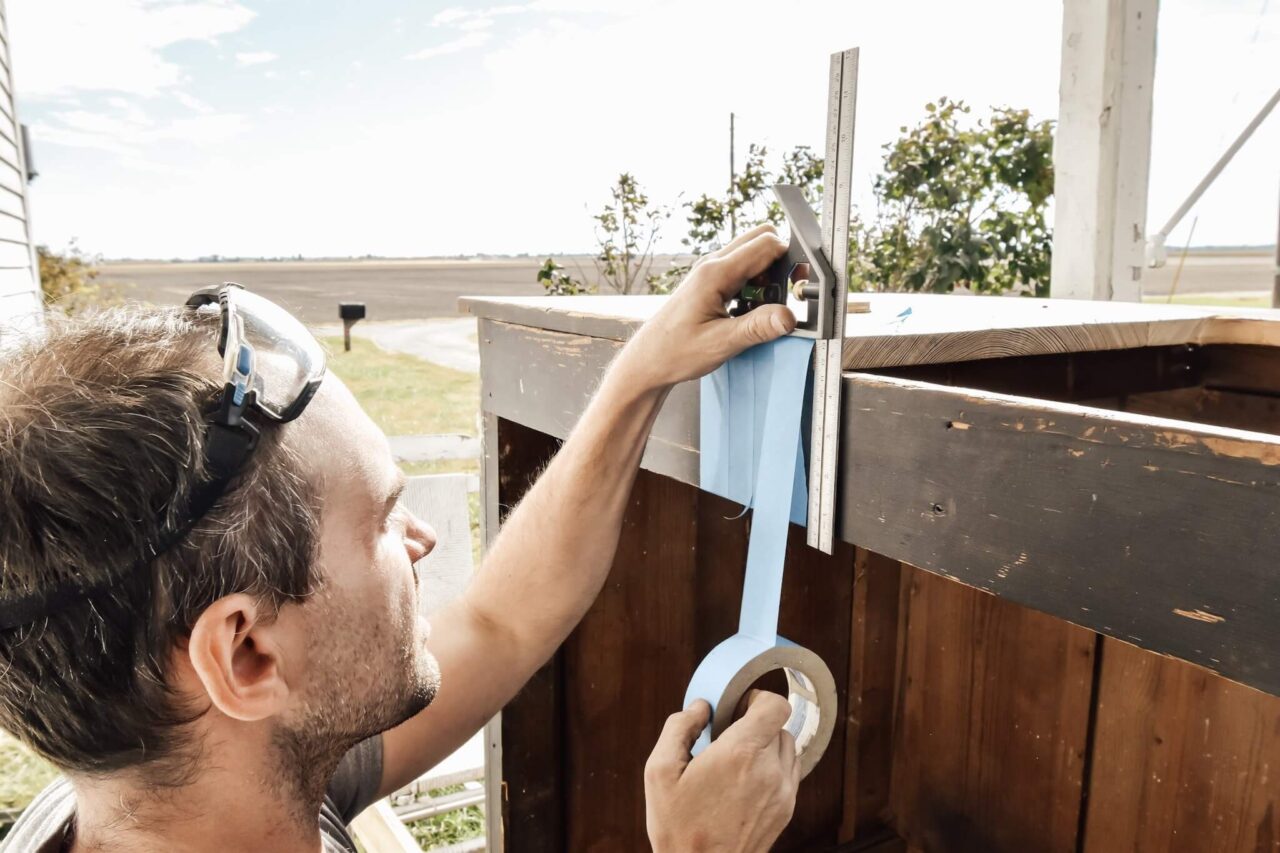 A man wearing safety goggles is applying blue painter's tape to a wooden surface using a square ruler for alignment.