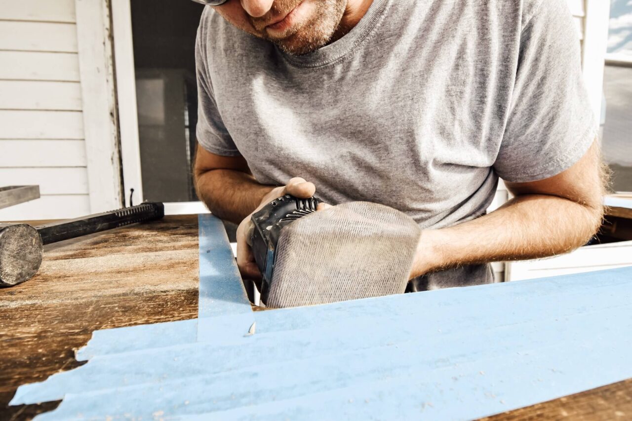 A man using a sander on a wooden surface that has blue painter's tape applied to it.