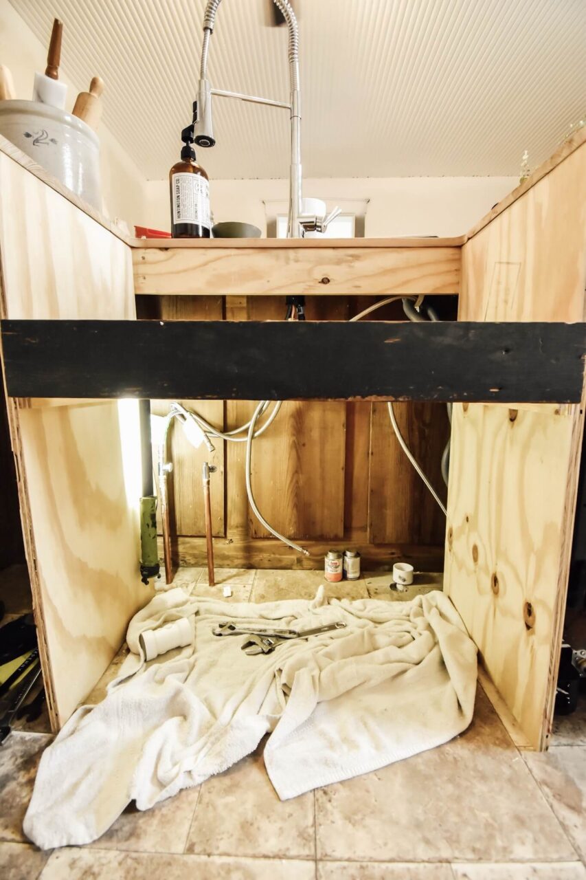 An under-sink area with exposed pipes and tools, with a towel spread on the floor and a modern faucet installed above.