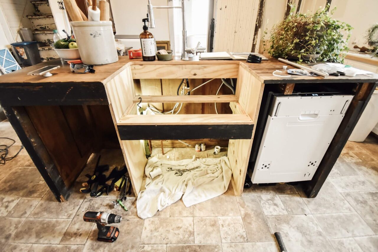 A kitchen setup under construction with a newly installed farmhouse sink, plumbing exposed beneath, and various tools and materials scattered around.