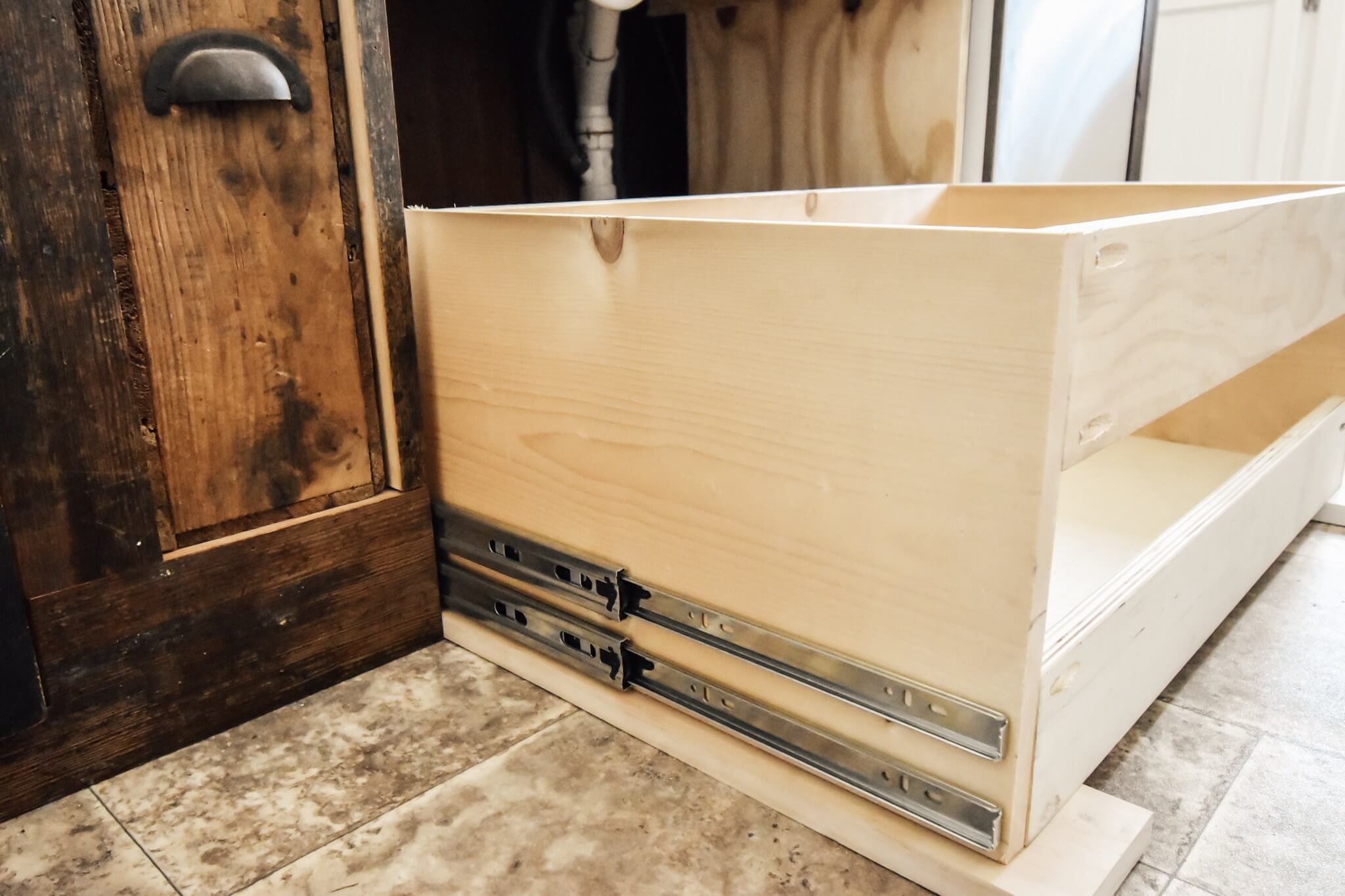 A partially assembled wooden drawer with metal slides installed, next to a rustic wooden cabinet in a kitchen.