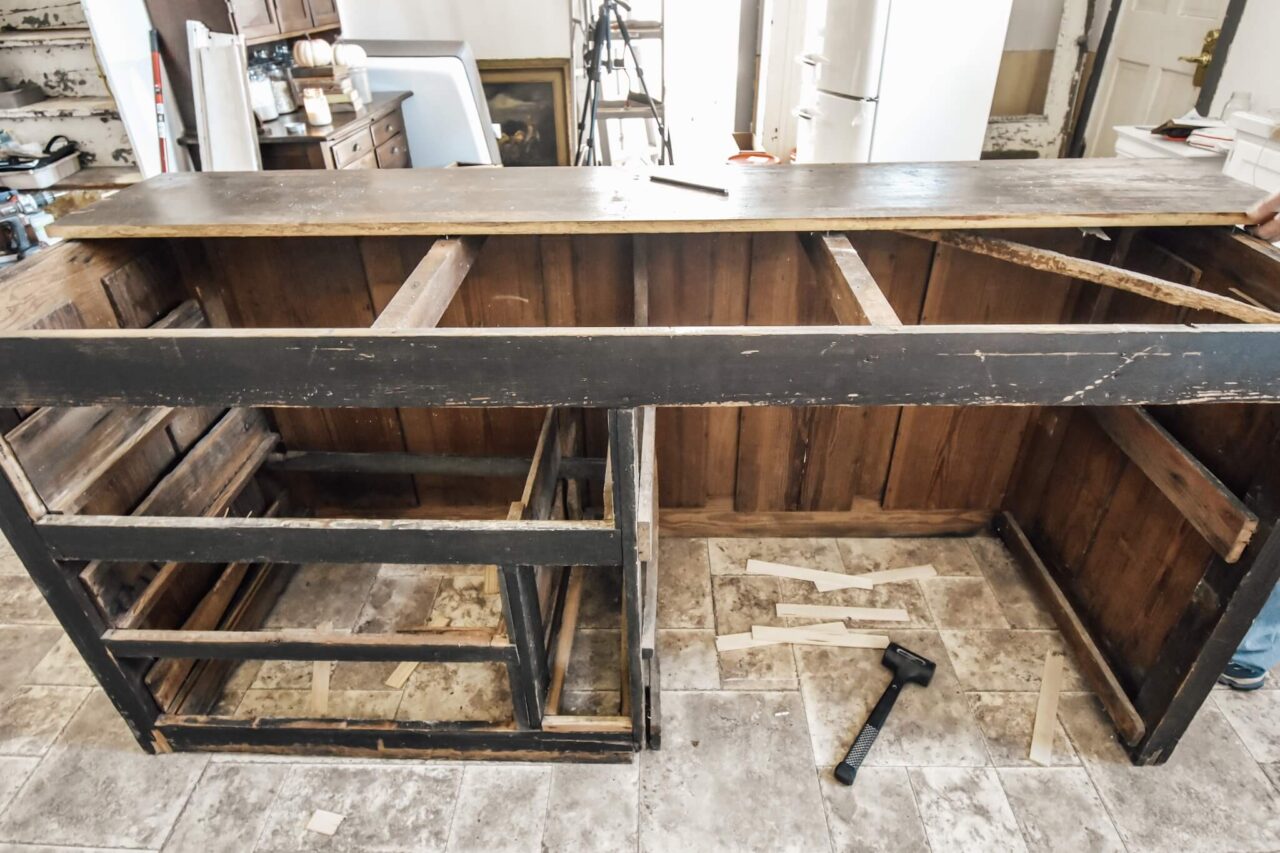 A partially disassembled rustic wooden kitchen island with its drawers and panels removed, placed in a room with tiled flooring and various tools on the ground.