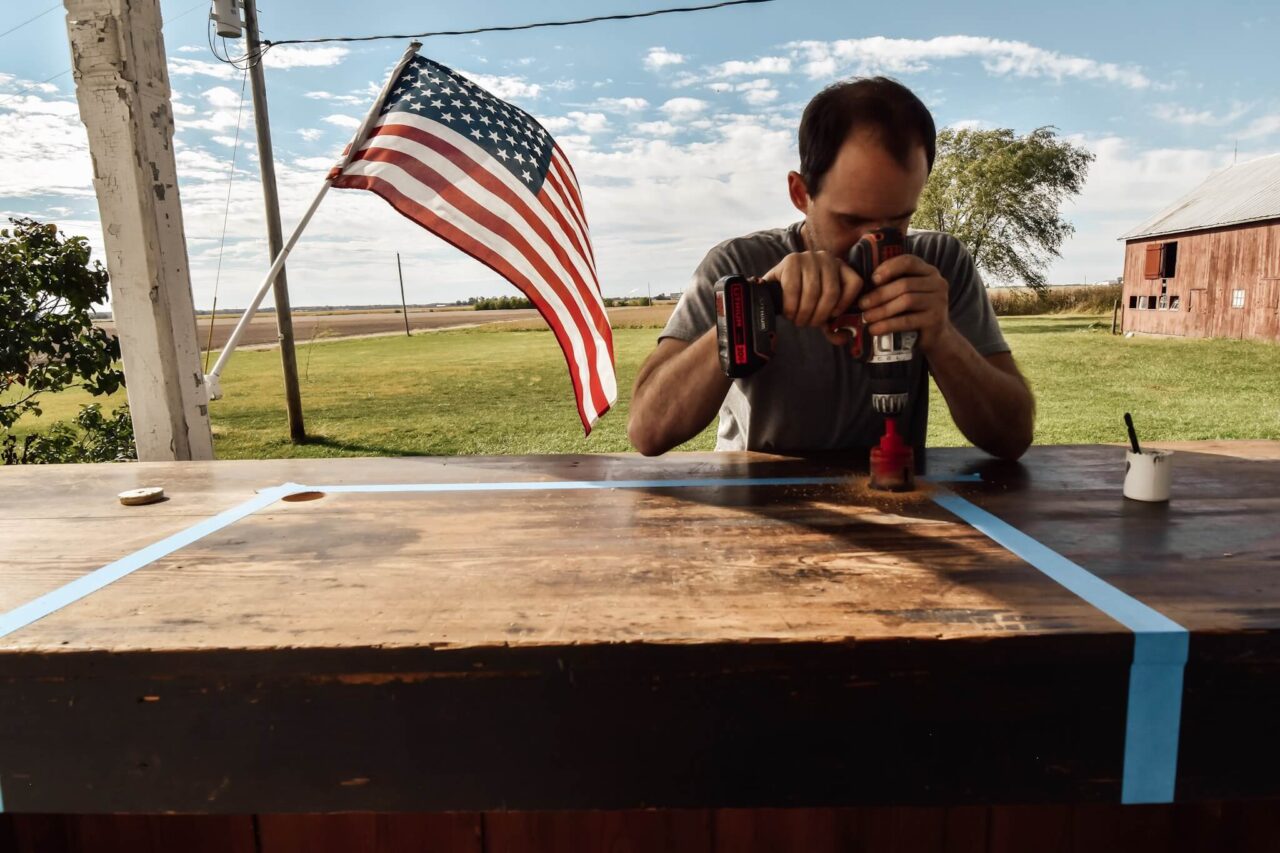 A man is drilling into a wooden surface outside, with an American flag waving in the background.