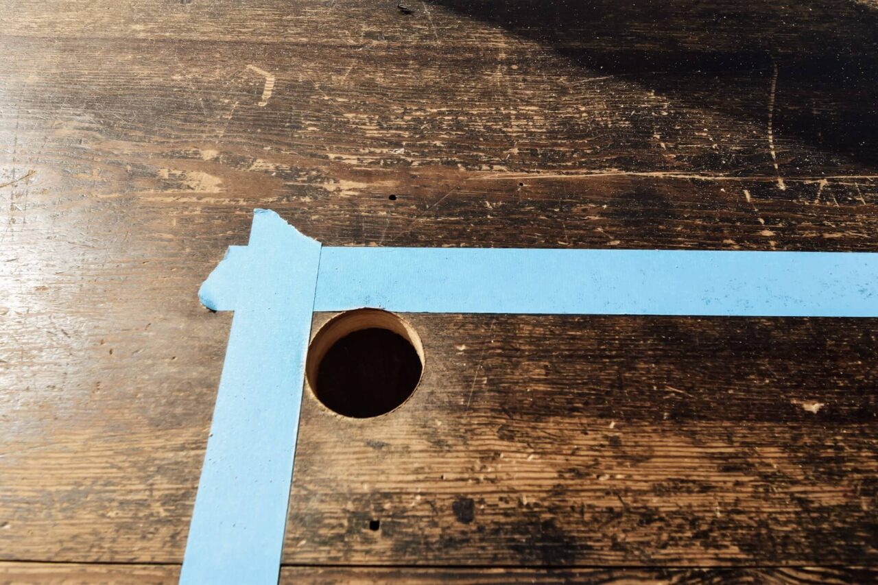 A close-up of a wooden surface with blue painter's tape forming a corner and a freshly drilled hole.