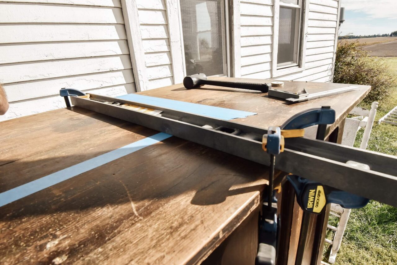 A wooden surface with blue painter's tape and a clamped straightedge, with tools and a white house in the background.