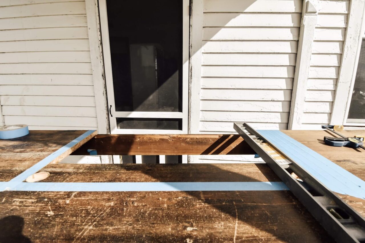 A wooden surface with a rectangular cutout marked by blue painter's tape, a clamped straightedge, and tools, in front of a white house.