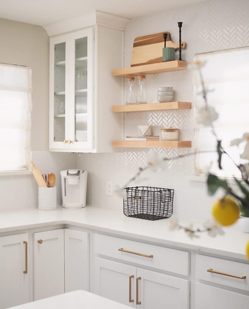 A bright kitchen with white cabinets, wooden floating shelves, and a countertop adorned with a black wire basket and a coffee maker.