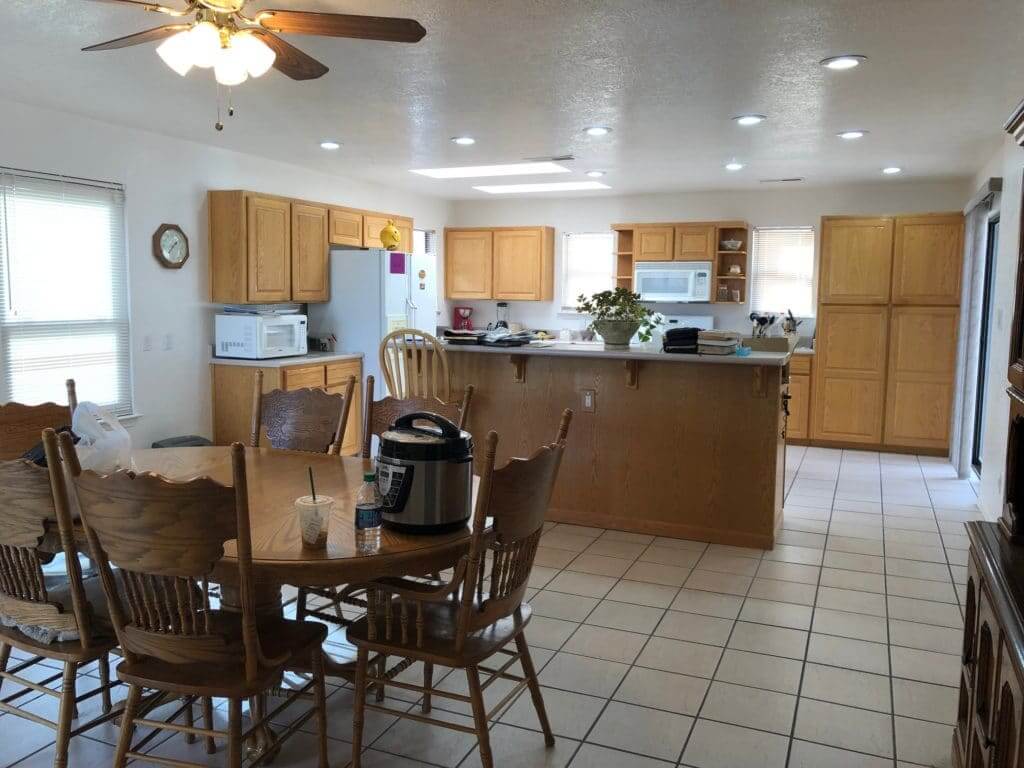 A kitchen with wooden cabinets, a dining table with chairs, and a ceiling fan, featuring a microwave, refrigerator, and various kitchen items on the counter.