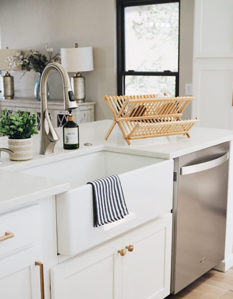 A kitchen sink with a striped towel draped over the front, a potted plant, soap dispenser, and wooden dish rack on the countertop, and a stainless steel dishwasher beneath.