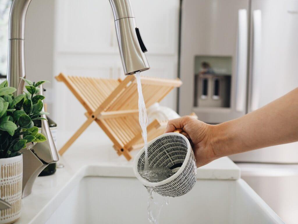 A hand rinsing a strainer under a kitchen faucet, with a potted plant and a wooden dish rack in the background.
