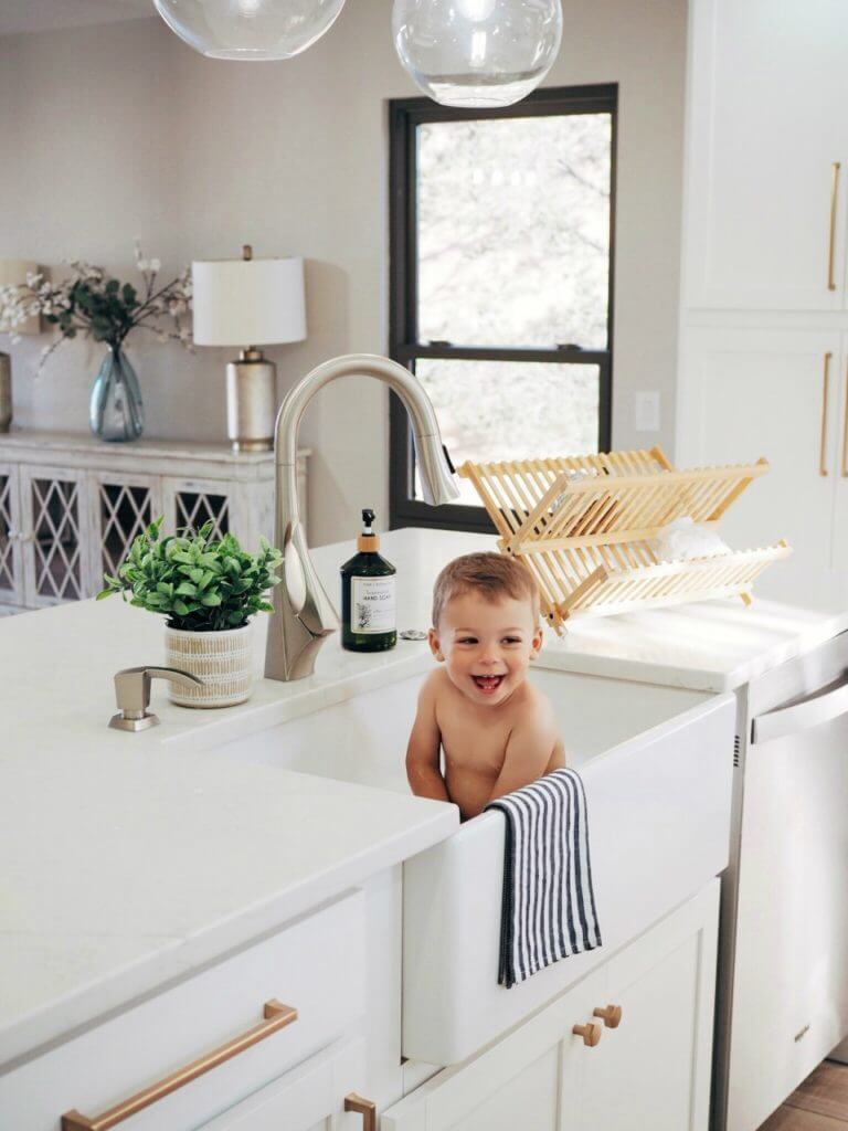 A baby sitting in a kitchen sink, smiling, with a striped towel hanging over the edge, a potted plant, and a wooden dish rack in the background.