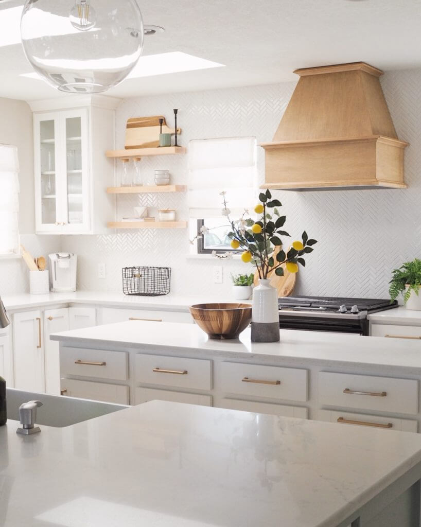 A modern kitchen with white cabinets, a wooden range hood, floating shelves, and a central island with a decorative bowl and vase with flowers.
