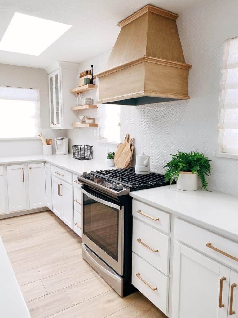 A bright kitchen with white cabinets, a wooden range hood, stainless steel oven, and open shelves displaying kitchen items, along with potted plants for decor.
