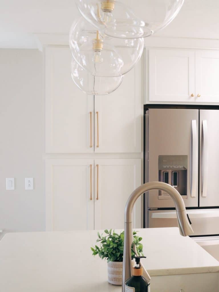 A modern kitchen featuring a stainless steel refrigerator, white cabinets with gold handles, a countertop with a plant and soap dispenser, and glass pendant lights.