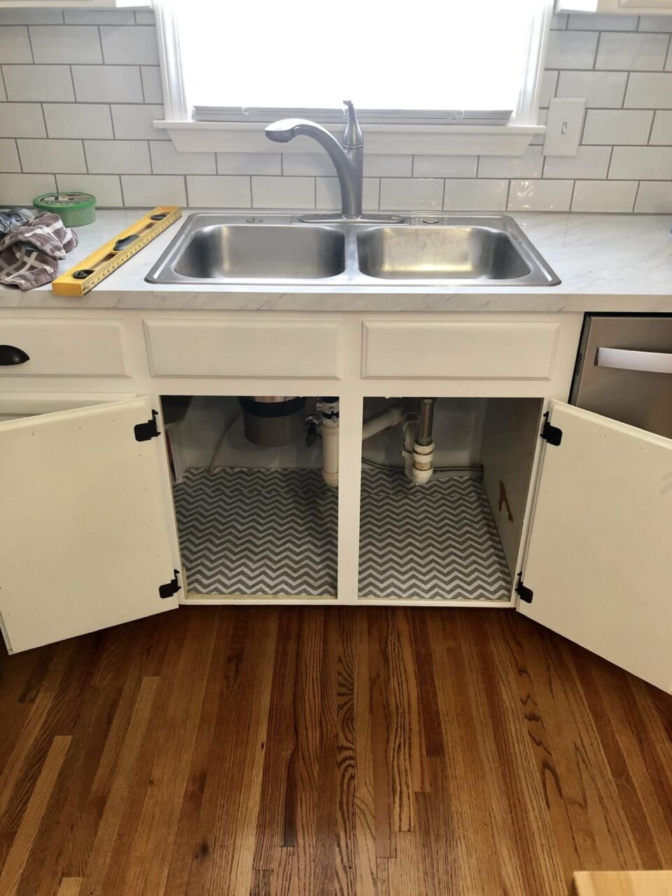 A kitchen sink with open cabinets below, showing exposed plumbing and chevron-patterned shelf liner.