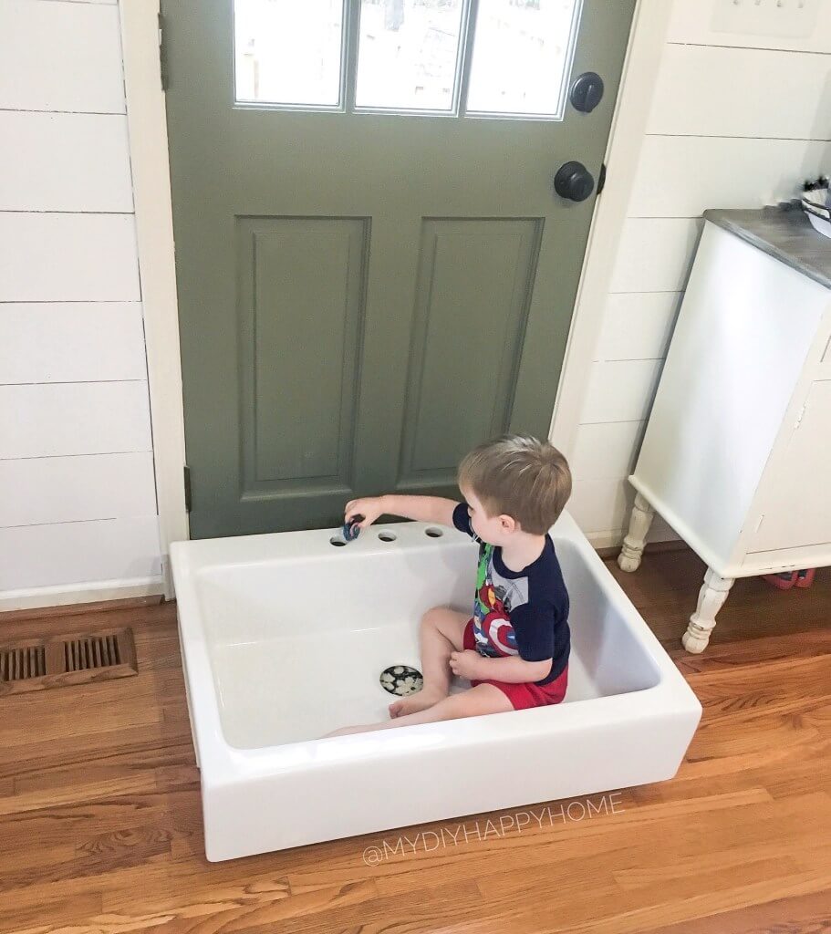 A young child sitting in a white basin near a green door, playing with a small toy car.