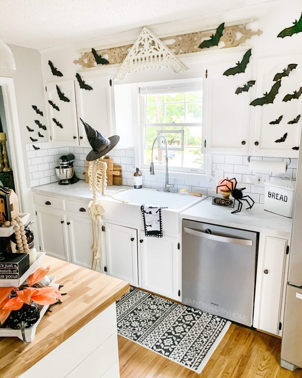 A kitchen decorated for Halloween with bat decals on the cabinets, a skeleton wearing a witch hat by the sink, and a spider decoration on the counter."